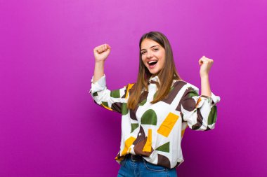 Genç ve güzel bir kadın muzaffer bir şekilde bağırıyor. Heyecanlı, mutlu ve şaşırmış görünüyor. Mor arka planda kutlama yapıyor.