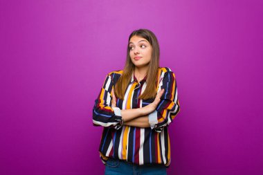 Genç, güzel bir kadın omuz silkiyor, kafası karışık ve kararsız, kolları bağlı ve şaşkın bir şekilde mor arka plana bakıyor.