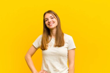 Genç ve güzel bir kadın mutlu bir şekilde gülümsüyor, eli kalçasında ve kendinden emin, pozitif, gururlu ve arkadaş canlısı bir tavırla turuncu arka plana karşı.