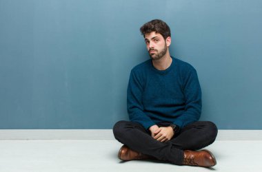 Genç yakışıklı bir adam üzgün ve mızmız bir şekilde yerde oturuyor. Mutsuz bir bakışla ağlıyor. Olumsuz ve hayal kırıklığına uğramış bir tavırla ağlıyor.