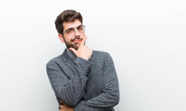 Genç yönetici ciddi, düşünceli ve güvensiz görünüyor, bir kolu çapraz ve çene üzerinde, seçenekleri beyaz duvara karşı tartıyor