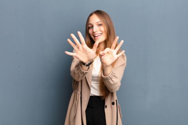 Genç ve güzel bir bayan gülümsüyor ve arkadaş canlısı görünüyor, 8 ya da 8 numarayı gösteriyor, elleri önde, gri arka planda geriye doğru sayıyor.