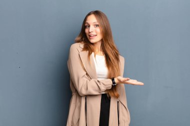 Genç, güzel bir kadın neşeyle gülümsüyor, mutlu hissediyor ve bir konsepti fotokopi uzayında gösteriyor, avuç içi gri arka planda.