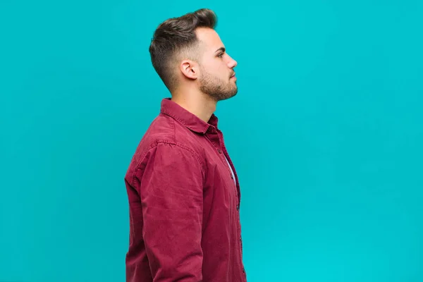 young hispanic man on profile view looking to copy space ahead, thinking, imagining or daydreaming against blue background