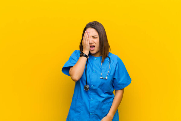 looking sleepy, bored and yawning, with a headache and one hand covering half the face isolated against yellow wall