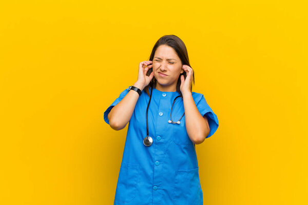 looking angry, stressed and annoyed, covering both ears to a deafening noise, sound or loud music isolated against yellow wall