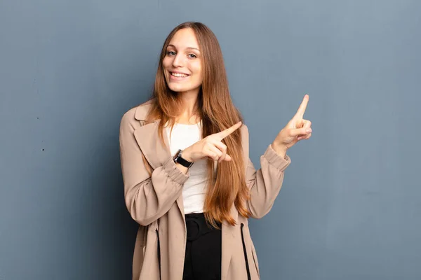 young pretty woman smiling happily and pointing to side and upwards with both hands showing object in copy space against gray background