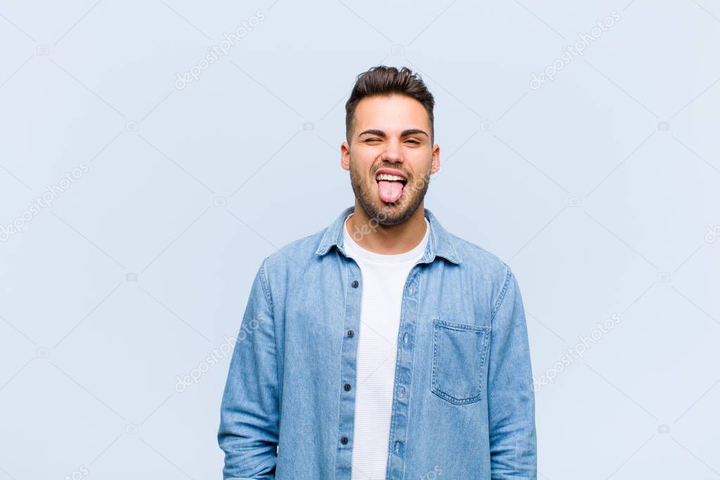 young hispanic man with cheerful, carefree, rebellious attitude, joking and sticking tongue out, having fun against blue wall