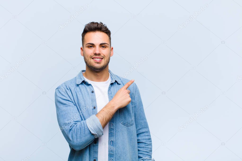 young hispanic man smiling cheerfully, feeling happy and pointing to the side and upwards, showing object in copy space against blue wall