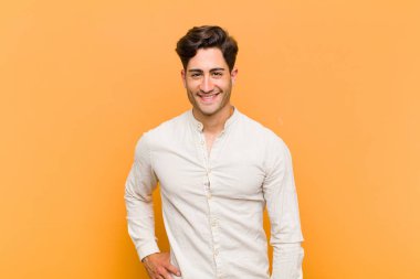 Genç yakışıklı adam mutlu bir şekilde gülümsüyor, eli kalçasında ve kendinden emin, pozitif, gururlu ve arkadaş canlısı bir tavırla turuncu arka plana karşı.