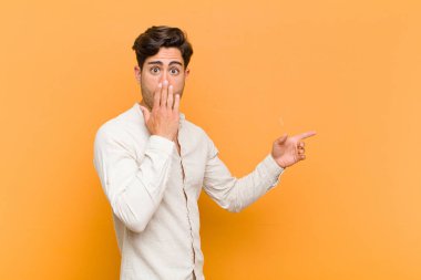 Genç yakışıklı adam kendini mutlu, şaşkın ve şaşırmış hissediyor. Ağzını eliyle kapatıyor ve turuncu arka plana karşı yanal fotokopi alanını işaret ediyor.