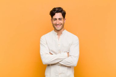 Genç yakışıklı adam kendini mutlu, gururlu ve umutlu hissediyor, merak ediyor veya düşünüyor, kollarını turuncu arka planda çapraz bağlamış bir şekilde fotokopi çekmek istiyor.