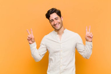Genç yakışıklı adam gülümsüyor ve mutlu görünüyor, arkadaş canlısı ve memnun, işaret eden zafer ya da barış her iki elinle turuncu arka planda