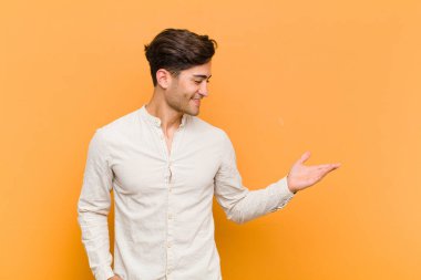 young handsome man feeling happy and cheerful, smiling and welcoming you, inviting you in with a friendly gesture against orange background