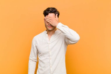 young handsome man covering eyes with one hand feeling scared or anxious, wondering or blindly waiting for a surprise against orange background
