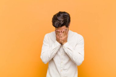 Genç yakışıklı adam üzgün, hayal kırıklığına uğramış, gergin ve depresif, iki eliyle yüzünü kapatıyor, turuncu arka planda ağlıyor.