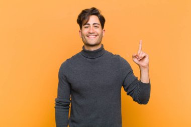 Genç yakışıklı adam mutlu ve mutlu bir şekilde gülümsüyor, bir eliyle turuncu arka plandaki boşluğu kopyalamak için yukarı bakıyor.