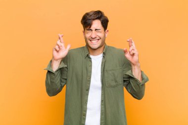 Genç yakışıklı adam gülümsüyor ve endişeyle iki parmağını çapraz yapıyor. Endişeli hissediyor ve turuncu arka planda iyi şanslar diliyor veya umut ediyor.