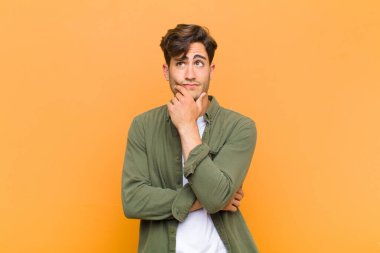 Genç yakışıklı düşünen, kuşkulu ve şaşkın hisseden, farklı seçenekleri olan, turuncu arka plana karşı hangi kararı vereceğini merak eden.