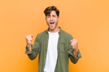 Kızgın bir ifadeyle ya da turuncu arka planda başarıyı kutlayan yumruklarla bağıran genç ve yakışıklı bir adam.