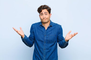 Genç yakışıklı adam şaşkın ve şaşkın, kuşkulu, farklı seçenekleri tartıyor ya da seçiyor mavi arkaplana karşı komik bir ifadeyle