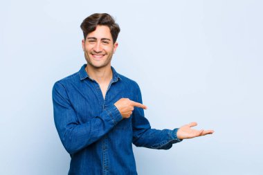 Genç yakışıklı adam gülümsüyor, mutlu, kaygısız ve tatmin olmuş hissediyor. Fotokopi alanındaki kavram ya da fikre işaret ediyor. Mavi arka planda.