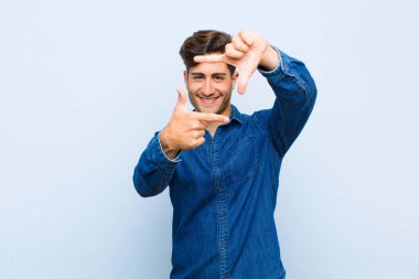 Genç yakışıklı adam mutlu, arkadaş canlısı ve pozitif hissediyor, gülümsüyor ve elleri mavi arka planda bir portre veya fotoğraf çerçevesi yapıyor.