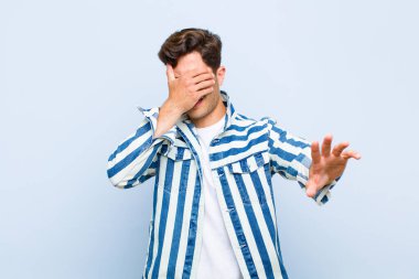 Genç ve yakışıklı bir adam yüzünü eliyle kapatıyor ve diğer elini kamerayı durdurmak için öne koyuyor. Mavi arka planda fotoğraf veya resimleri reddediyor.