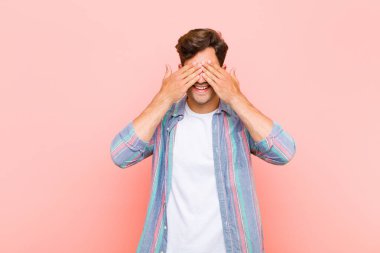 Genç yakışıklı adam gülümsüyor ve mutlu hissediyor, iki eliyle gözlerini kapatıyor ve pembe arka planda inanılmaz bir sürpriz bekliyor.