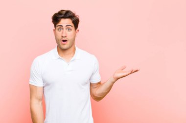 Genç yakışıklı adam şaşırmış ve şok olmuş görünüyor, çenesi düşmüş, pembe arka planda eli açık bir objeyi tutuyor.