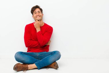 Genç yakışıklı adam gülümsüyor, hayattan zevk alıyor, mutlu, arkadaş canlısı, tatmin olmuş ve elleri çenesinde yerde oturuyor.