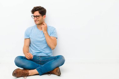 Genç ve yakışıklı bir adam stresli, hayal kırıklığına uğramış ve yorgun, ağrılı boynunu ovuyor, endişeli, sorunlu bir bakışla yerde oturuyor.