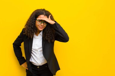 Siyah iş kadını şaşkın ve şaşkın görünüyor, el üstünde uzak bakıyor, turuncu duvarın karşısında izliyor ya da arıyor