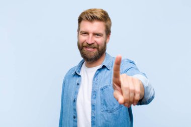 Genç, sarışın yetişkin bir adam gururla gülümsüyor ve gururla bir numaralı pozunu veriyor, kendini lider gibi hissediyor.