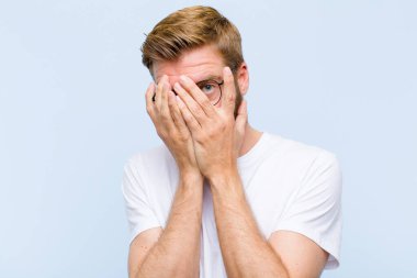 Genç, sarışın, yetişkin bir adam korkmuş ya da utanmış hissediyor. Gözleri yarı kapalı ya da ellerle dikizliyor ya da dikizliyor.