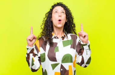 Genç, güzel bir kadın şaşırmış, şaşırmış ve ağzı açık görünüyor, iki eliyle yukarı doğru uzayı kroma tuşlu duvara kopyalamak için işaret ediyor.