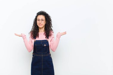 Genç güzel kadın şaşkın ve şaşkın, şüpheci, farklı seçenekleri tartıyor ya da beyaz duvara karşı komik bir ifadeyle seçiyor.