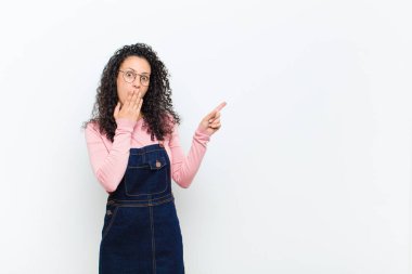 Genç, güzel bir kadın kendini mutlu, şok olmuş ve şaşırmış hissediyor. Ağzı eliyle kapatıyor ve beyaz duvara karşı yanal fotokopi alanını işaret ediyor.