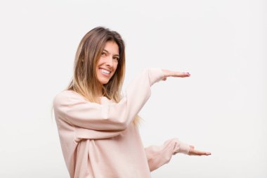 Genç, güzel bir kadın gülümsüyor, mutlu, pozitif ve tatmin olmuş hissediyor, beyaz arkaplana karşı fotokopi alanında nesne ya da konsept tutuyor ya da gösteriyor