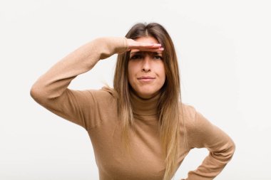 Genç ve güzel bir kadın şaşkınlık ve şaşkınlık içinde bakıyor, uzaktan bakıyor, beyaz arkaplanı izliyor ya da araştırıyor.