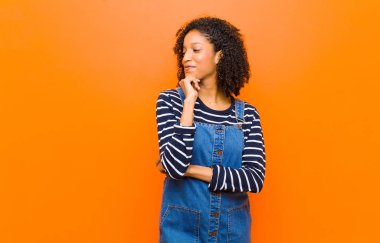 Genç, güzel, siyah bir kadın mutlu, kendinden emin bir ifadeyle gülümsüyor. Elleri çenesinde, turuncu duvara bakıyor ve merak ediyor.