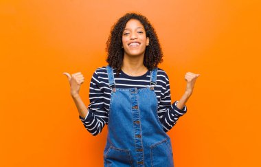 Genç ve güzel siyahi kadın neşeyle gülümsüyor ve mutlu görünüyor, kaygısız ve pozitif hissediyor, iki başparmağı turuncu duvara dayalı.