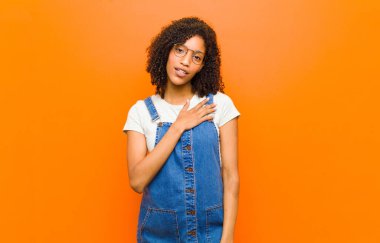 Genç ve güzel siyah kadın şok olmuş ve şaşırmış hissediyor, gülümsüyor, el ele tutuşuyor, mutlu ya da turuncu duvara karşı minnettarlık gösteriyor.