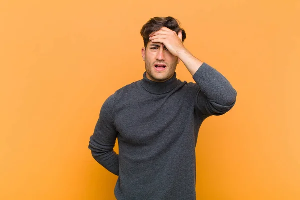 young handsome man panicking over a forgotten deadline, feeling stressed, having to cover up a mess or mistake against orange background