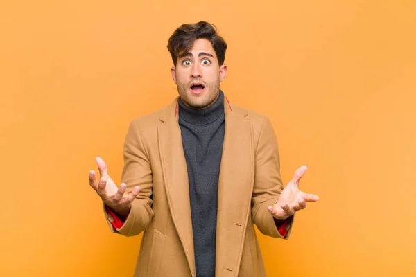 young handsome man open-mouthed and amazed, shocked and astonished with an unbelievable surprise against orange background