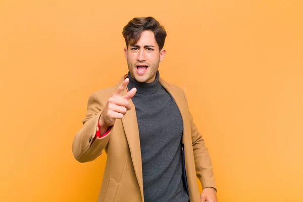 young handsome man pointing at camera with an angry aggressive expression looking like a furious, crazy boss against orange background