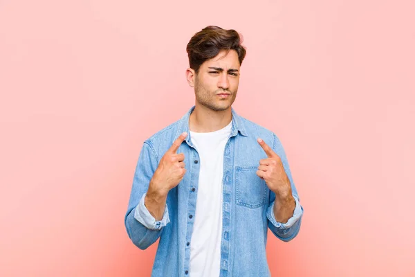 young handsome man with a bad attitude looking proud and aggressive, pointing upwards or making fun sign with hands against pink background