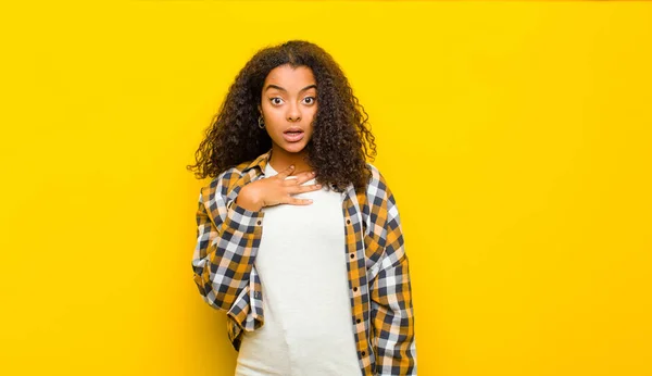 young pretty african american woman feeling shocked, astonished and surprised, with hand on chest and open mouth, saying who, me? against yellow wall