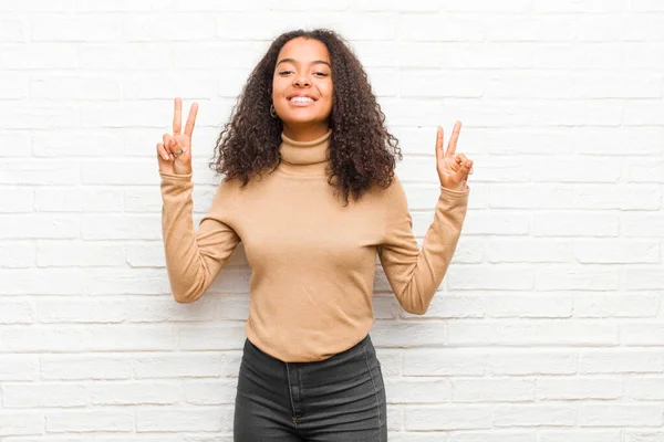 young black woman smiling and looking happy, friendly and satisfied, gesturing victory or peace with both hands against brick wall
