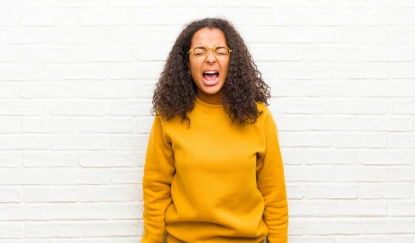 young black woman shouting aggressively, looking very angry, frustrated, outraged or annoyed, screaming no against brick wall